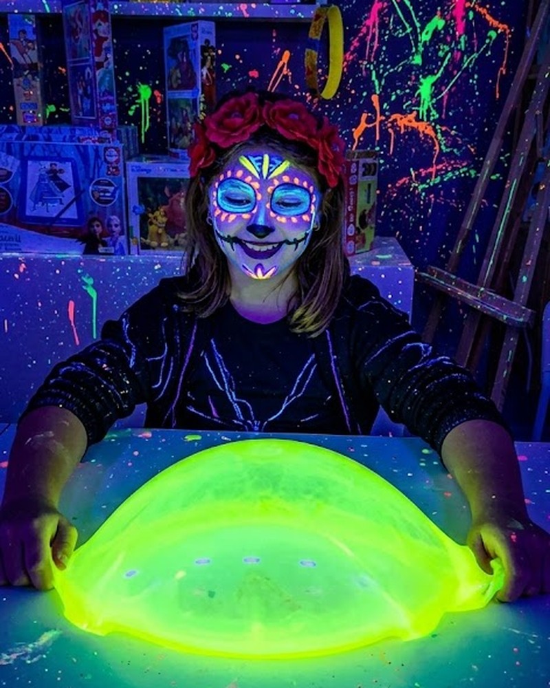 Niña con la cara pintada de catrina fluorescente estirando una gran bola de slime amarillo neón en la sala de luz ultravioleta